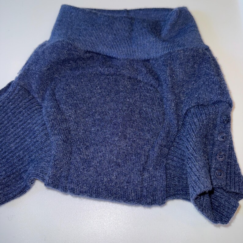 May include: A blue sweater with a wide, folded collar. The sweater has long sleeves and buttons on the cuff. The fabric appears to be a soft knit.