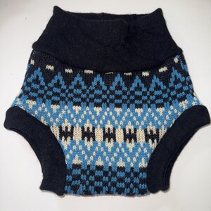 May include: A pair of knitted nappy covers featuring a geometric pattern in shades of black, bright blue, and cream. The garment has a wide, solid black ribbed waistband and solid black ribbed leg openings, constructed with a soft, textured knit fabric.