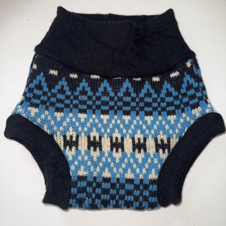 May include: A pair of knitted nappy covers featuring a geometric pattern in shades of black, bright blue, and cream. The garment has a wide, solid black ribbed waistband and solid black ribbed leg openings, constructed with a soft, textured knit fabric.
