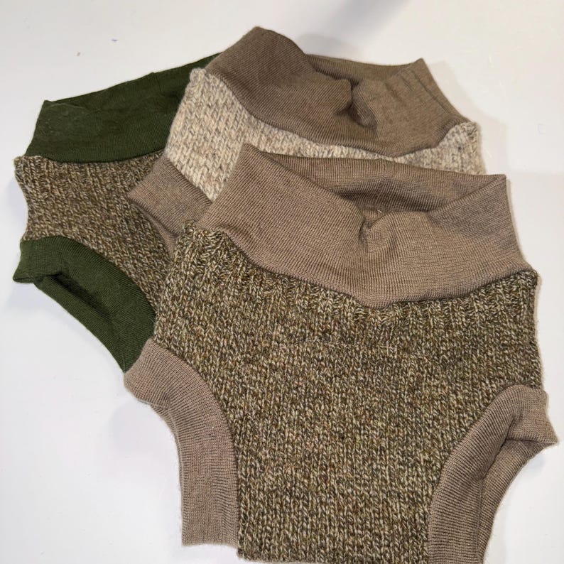 May include: Three pairs of knitted baby shorts in shades of green, brown, and beige. The shorts feature a high-waisted design with ribbed cuffs and waistbands. The fabric has a textured appearance, suggesting a soft, comfortable material.