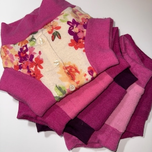 Puede incluir: Una pila de suéteres para bebés rosas y magenta. Un suéter tiene un estampado floral en la parte delantera con tres botones pequeños. Los otros suéteres son de colores lisos, que van desde el rosa claro hasta el magenta oscuro.