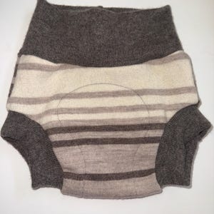 May include: A knitted diaper cover in shades of brown and cream. The cover features horizontal stripes and a high waistband. The leg openings and waistband are a darker brown. The cover is designed to be worn over a nappy.