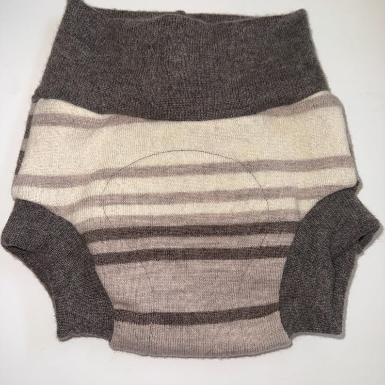 May include: A knitted diaper cover in shades of brown and cream. The cover features horizontal stripes and a high waistband. The leg openings and waistband are a darker brown. The cover is designed to be worn over a nappy.