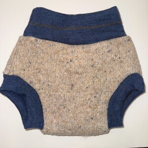 May include: A knitted diaper cover in a neutral speckled beige with navy blue trim. The waist and leg openings are navy blue. A thin brown stripe accents the navy blue waistband. The diaper cover is made of a soft, textured material.