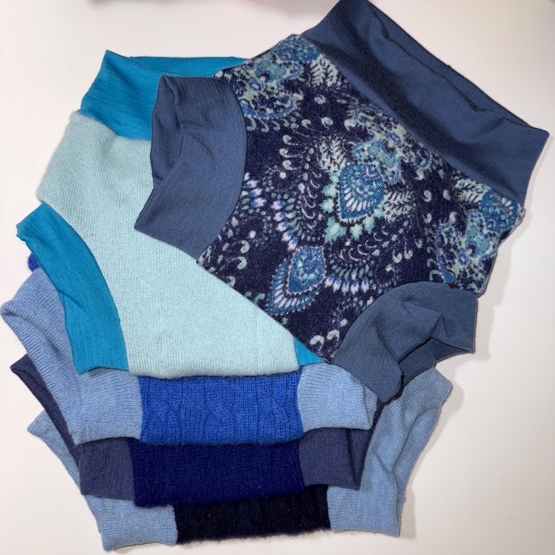 May include: A stack of various blue turtleneck sweaters. The sweaters range in shades from light blue to navy, with one featuring a patterned design. The sweaters are made of different materials, including knit and patterned fabric.
