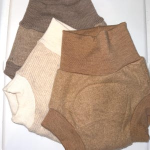 May include: Three pairs of baby bloomers in shades of beige and brown are displayed. The bloomers have a high-waisted design with ribbed cuffs. The colours vary from a light cream to a rich, warm brown. The fabric has a soft, textured appearance.