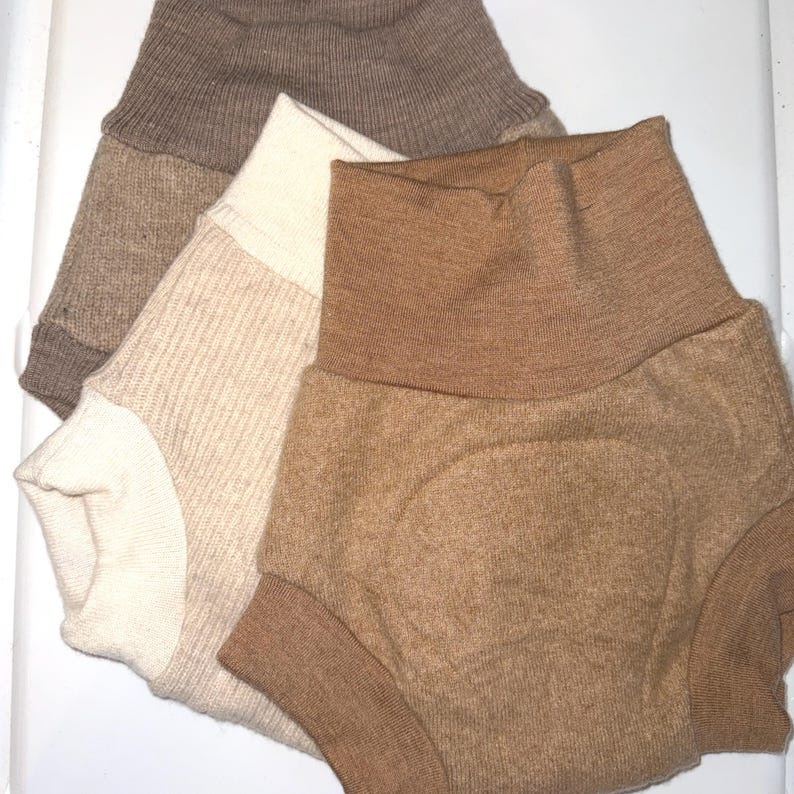 May include: Three pairs of baby bloomers in shades of beige and brown are displayed. The bloomers have a high-waisted design with ribbed cuffs. The colours vary from a light cream to a rich, warm brown. The fabric has a soft, textured appearance.