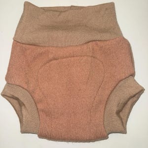 May include: Knitted baby bloomers in peach with a tan waistband and cuffs. The high-waisted bloomers have a rounded shape. The garment is designed for a baby.