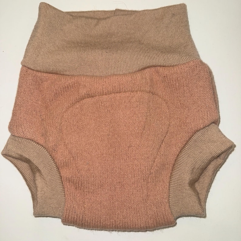 May include: Knitted baby bloomers in peach with a tan waistband and cuffs. The high-waisted bloomers have a rounded shape. The garment is designed for a baby.