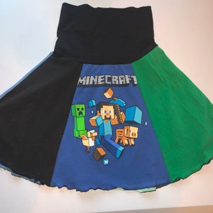 May include: A black, green, and blue Minecraft-themed skirt. The skirt features a black waistband and panels of black and green fabric. The front panel is blue with the Minecraft logo and character graphics.