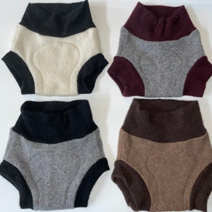 Puede incluir: Se muestran cuatro pañales de tela en varios colores. Los pañales tienen una cintura alta y están hechos de tela suave. Los colores incluyen negro, crema, gris y marrón. Cada pañal tiene un ribete de color contrastante alrededor de las piernas y la cintura.