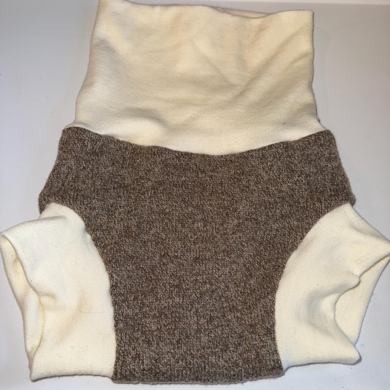May include: A two-tone baby nappy cover. The top is a cream-coloured, ribbed fabric that folds over. The bottom is a brown, knitted fabric with cream-coloured leg openings. The nappy cover is designed to be soft and comfortable.