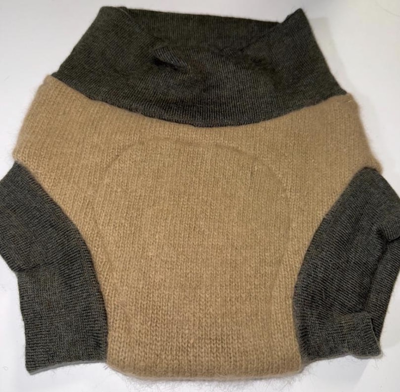 May include: A two-tone sweater in a neutral colour palette. The upper section and sleeves are a dark olive green, while the body is a light tan. The sweater has a high, folded collar and long sleeves. The knit fabric appears soft and warm.