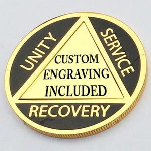 May include: Gold-colored coin with a black triangle in the center. The triangle has the words "Unity", "Service", and "Recovery" on each point. The words "Custom Engraving Included" are printed inside the triangle.