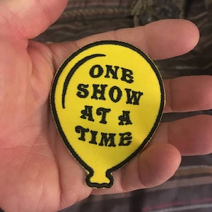May include: Yellow balloon patch with black outline and text that reads "One Show At A Time".