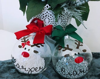 May include: Two clear glass Christmas baubles decorated with reindeer faces and filled with white and silver glitter. The baubles are personalised with the names "Harper" and "Cecilia".