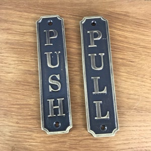 Könnte beinhalten: Zwei antike goldfarbene Metallschildchen mit schwarzem Hintergrund. Die Schilder sind rechteckig und haben abgerundete Ecken. Das linke Schild zeigt "PUSH" in großen, erhabenen Buchstaben. Das rechte Schild zeigt "PULL" in großen, erhabenen Buchstaben.