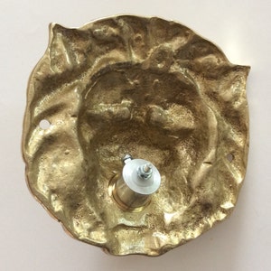 May include: Brass door knocker with a detailed design, featuring a face with a textured surface. The knocker is attached to a metal mounting plate with a screw.