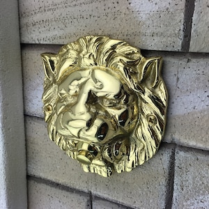 Puede incluir: Un llamador de puerta de cabeza de león de oro con un diseño detallado. El llamador tiene un botón redondo en el centro de la boca.