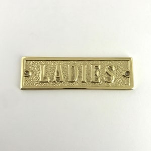 May include: A gold-colored metal sign with the word "LADIES" embossed on it.