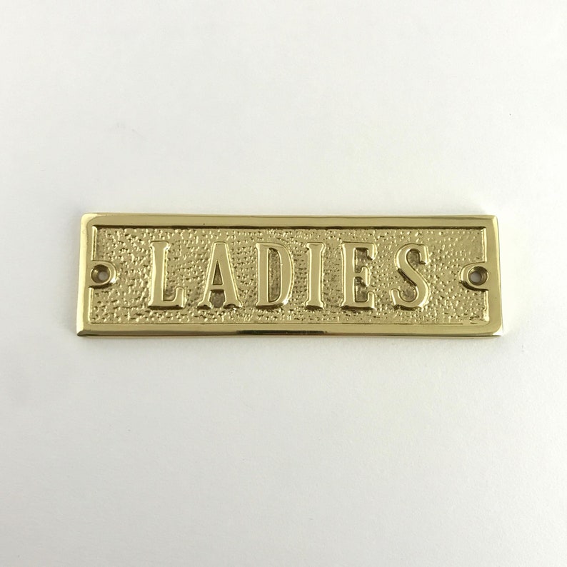 May include: A gold-colored metal sign with the word "LADIES" embossed on it.