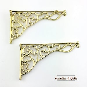 Op de afbeelding: Twee gouden metalen plankbeugels met een decoratief, sierlijk, scroll-ontwerp. De beugels zijn in de vorm van een rechte hoek.