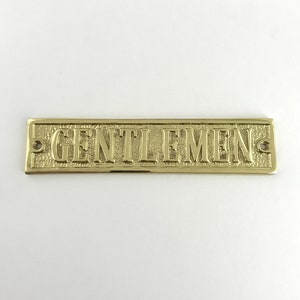 May include: A gold-colored brass sign with the word "GENTLEMEN" embossed on it.