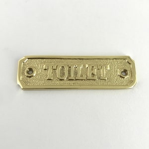 May include: A gold-colored metal sign with the word "TOILET" embossed on it. The sign has two small holes on either side for hanging.