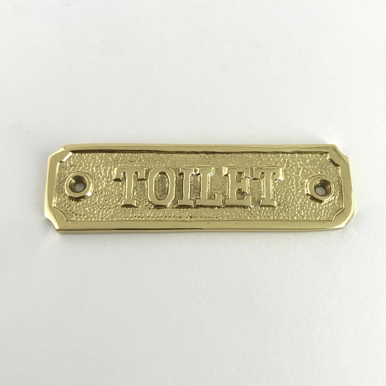 May include: A gold-colored metal sign with the word "TOILET" embossed on it. The sign has two small holes on either side for hanging.