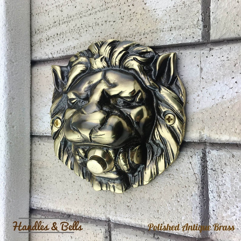 May include: A polished antique brass lion head doorbell. The doorbell is mounted on a white brick wall. The lion's mouth is open and the doorbell button is visible.