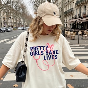 May include: Ivory-colored t-shirt with the text "PRETTY GIRLS SAVE LIVES" in blue and pink, featuring a stethoscope design. The model is wearing a beige baseball cap with a black logo and a black purse with a chain strap.