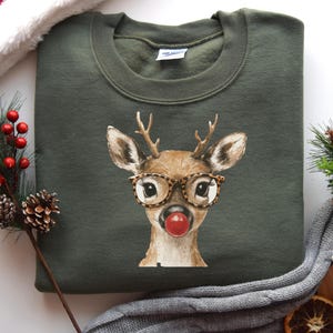 Può includere: Una felpa verde oliva con una grafica di cervo stravagante. Il cervo indossa occhiali con stampa leopardata e un naso rosso. La felpa è circondata da decorazioni natalizie, tra cui un cappello di Babbo Natale e pigne.
