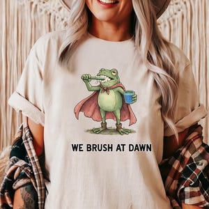 May include: Beige t-shirt featuring a cartoon frog brushing its teeth, wearing a cape and holding a blue cup. The text "WE BRUSH AT DAWN" is below the frog. The word "NATURAL" is printed at the bottom right.