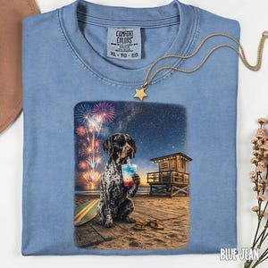 Peut inclure: Un t-shirt bleu avec une étiquette Comfort Colors présentant un motif de chien tacheté portant des lunettes de soleil, tenant une boisson rouge, blanche et bleue sur une plage la nuit avec des feux d'artifice, un poste de secours, une planche de surf et des sandales.