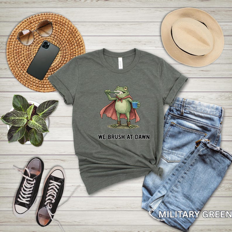 May include: Military green t-shirt featuring a cartoon frog superhero brushing its teeth, holding a blue cup, and wearing a red cape. The text "WE BRUSH AT DAWN" is below the frog. The shirt is paired with blue jeans, a straw hat, and black sneakers.