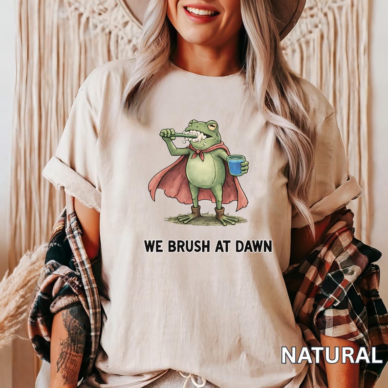 May include: Beige t-shirt featuring a cartoon frog brushing its teeth, wearing a cape and holding a blue cup. The text "WE BRUSH AT DAWN" is below the frog. The word "NATURAL" is printed at the bottom right.