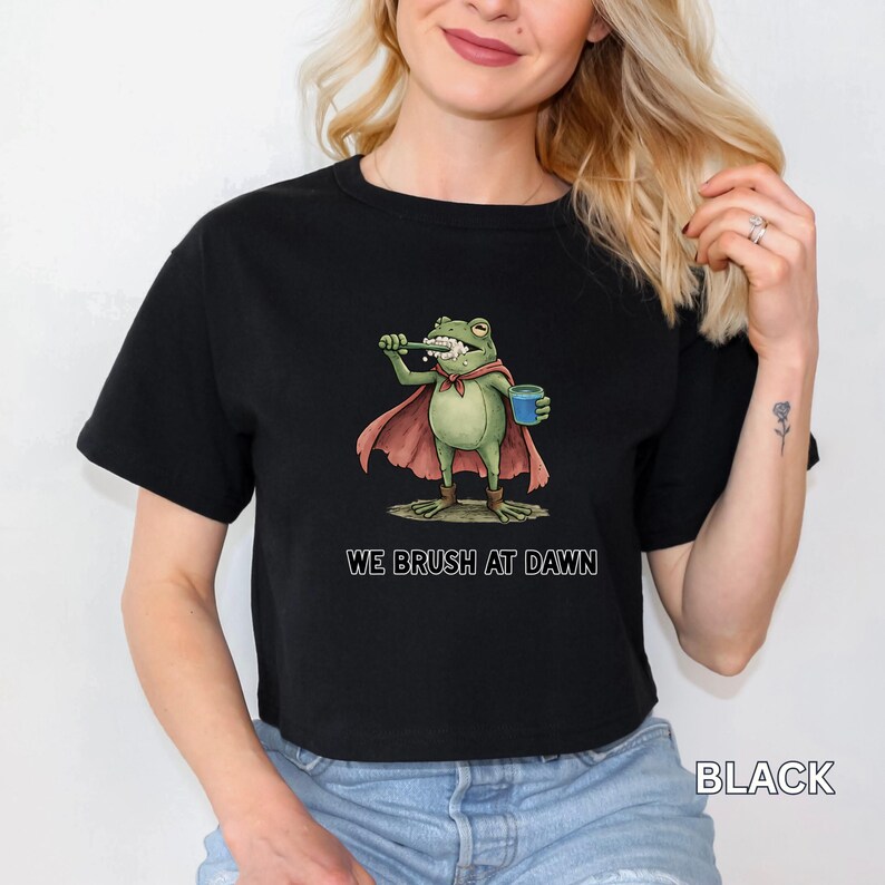 May include: Black t-shirt featuring a cartoon frog brushing its teeth, wearing a cape and holding a blue cup. The text "WE BRUSH AT DAWN" is printed below the frog. The word "BLACK" is printed at the bottom right.