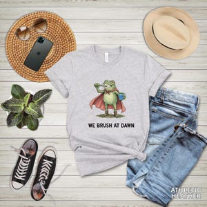 May include: Athletic heather gray t-shirt featuring a cartoon frog superhero brushing its teeth, wearing a red cape and holding a blue cup. The text "WE BRUSH AT DAWN" is printed below the frog. The shirt is paired with blue jeans and black sneakers.