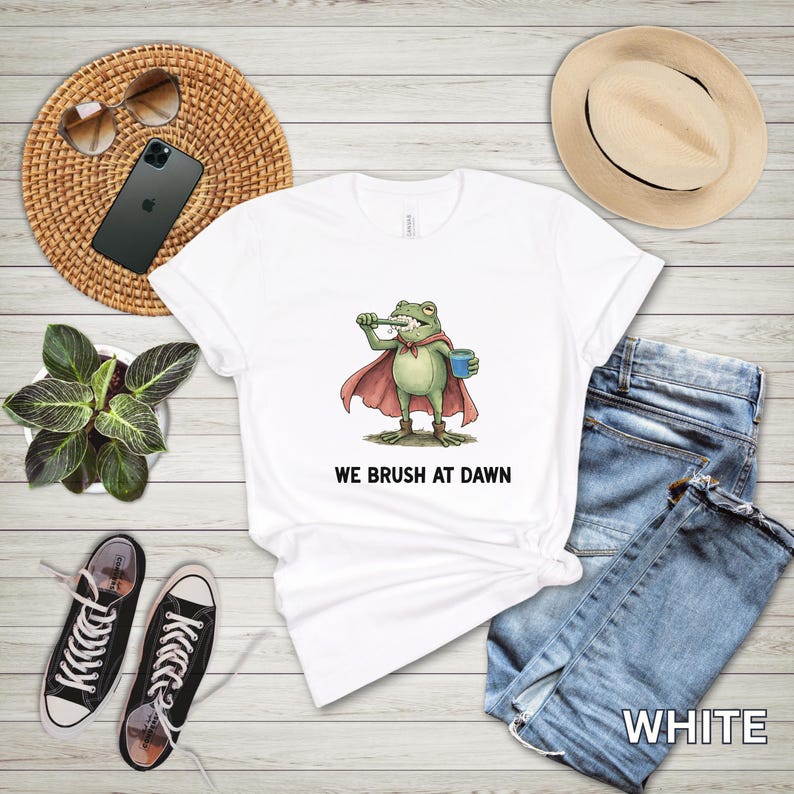May include: White t-shirt featuring a cartoon frog superhero brushing its teeth, with a red cape and blue cup. The text "WE BRUSH AT DAWN" is printed below the frog. The shirt is paired with blue jeans, a straw hat, and black sneakers.