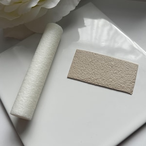 May include: A close-up of a white textured foam roller and a beige textured rectangle of material. Both items are on a white surface.