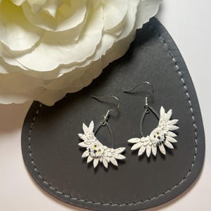 May include: A pair of white, floral earrings with silver hooks. The earrings are displayed on a dark grey, teardrop-shaped surface, with a white flower in the background. The earrings have a unique, handcrafted appearance.