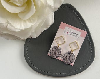 May include: A pair of dangle earrings with a floral pattern. The earrings have a gold square top and a round bottom with a black and white floral design. The earrings are displayed on a gray, heart-shaped tray with a white flower in the background. The earrings are from NALO uk Polymer Clay.