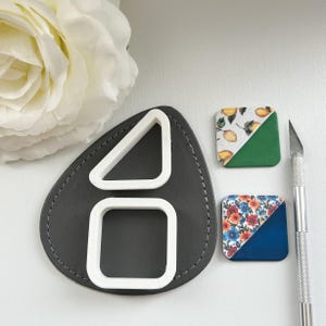 May include: A craft supplies image featuring a gray leather shape with white geometric cutters: a triangle and a square. Two square tiles with floral and lemon patterns are also present, along with a silver craft knife. The tiles have a diagonal color split.