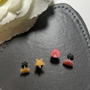 May include: Three pairs of earrings with a beach theme. The earrings are made of resin and feature a seashell, a starfish, and a seashell with a wave design. The earrings are all in shades of pink and yellow.