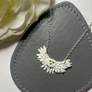 May include: A silver chain necklace with a white owl pendant. The owl has black eyes and yellow accents. The owl's wings are spread out and made of white feathers.