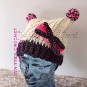 May include: A white and gray crocheted hat with a pink bow and two pom-poms on top. The hat is on a blue mannequin head. "Written Crochet Pattern" is written on the left side of the image.