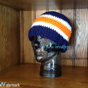 Op de afbeelding: Een donkerblauwe gebreide beanie met witte en oranje strepen. De beanie zit op een mannequinhoofd.