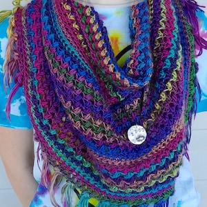 May include: A colorful crochet scarf with fringe. The scarf is made with a variety of colors, including purple, blue, green, and pink. The scarf is worn by a person, and a silver pendant hangs from the scarf.