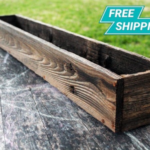 May include: A long, rectangular wooden planter box with a dark brown stain. The planter is made of reclaimed wood and has a rustic look.
