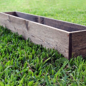 May include: A long, rectangular wooden planter box with a dark brown stain. The planter is sitting on a bed of green grass.